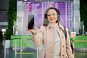 Pretty woman showing her mobile phone with blank screen to camera, standing by flight information panel and check-in board in