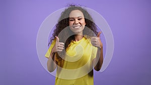 Pretty woman showing thumb up like sign over violet studio background. Positive young girl with curly hair smiles to