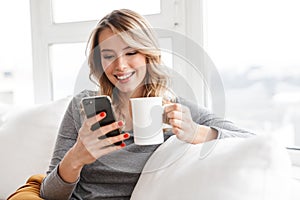 Pretty woman sitting indoors at home holding cup drinking tea using mobile phone