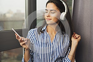 Pretty woman sitting by window and listening to relaxing music in headphones. Stream favorite songs directly from social networks