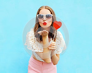 Pretty woman in sunglasses with red heart lollipop sends an air kiss over colorful blue