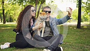 Pretty woman is taking selfie with her boyfriend and adorable dog using smartphone while resting in the park on the