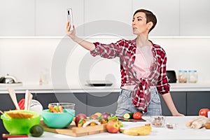 Pretty woman taking selfie or making a video call using her smartphone holding it in outstretched arm while cooking