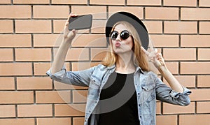 Pretty woman taking selfie picture by smartphone blowing kiss wearing a black round hat over brick background