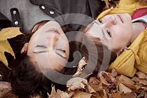 Pretty woman and teen girl are posing in autumn park. They are lying on fallen leaves. Beautiful landscape at fall season