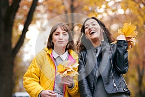 Pretty woman and teen girl are posing with bunch of maple`s leaves in autumn park. They are training for kiss. Beautiful landscap