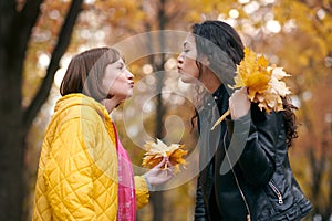Pretty woman and teen girl are posing with bunch of maple`s leaves in autumn park. They are training for kiss. Beautiful landscap