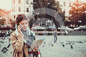 Pretty woman tourist talking on mobile phone while looking at map of the city, surrounded by flying pigeons on a square.