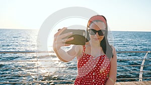 Pretty woman using black mobile phone by the sea beach. Girl in retro red polka dots dress making selfie, self portrait