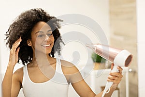 Pretty woman is using a hair dryer and smiling while looking in the mirror.
