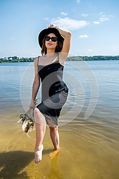 Pretty woman walking along barefoot in evening black dress in city lake