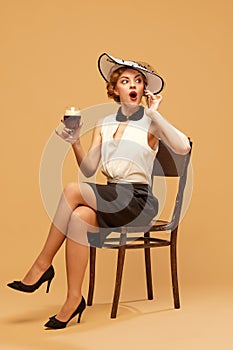 Pretty woman wearing elegant clothes with hat holding glass of beer and talking via cell phone over beige background