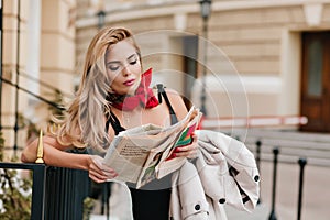 Pretty woman wears golden pendant reading newspaper while waiting friend on the street. Lovely concentrated lady looking