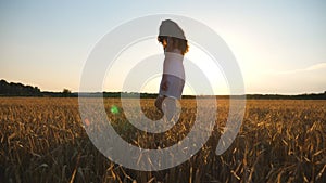 Pretty woman in white dress walking through field of yellow wheat with sunset at background. Young carefree girl