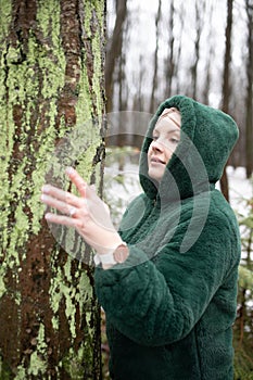 Pretty woman in winter forest. Female portrait in snow park. Woman in hood of eco fur jacket touch with hand tree trunk.