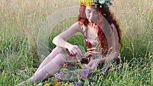 Pretty woman in wreath of sits in dry grass and touches wild flowers at summer