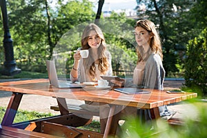 Pretty women sitting outdoors in park drinking coffee using laptop