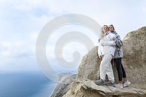 Pretty women stand on a high mountain on the sea coast on a sunny day. Smiling blondes hugging. Active lifestyle and recreation.