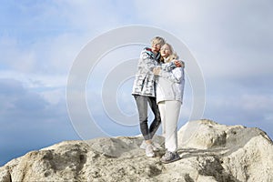 Pretty women stand on a high mountain on the sea coast on a sunny day. Smiling blondes hugging. Active lifestyle and recreation.