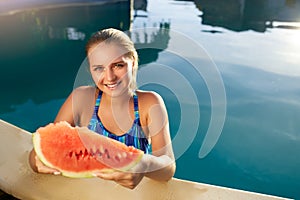 Pretty tanned slim young woman holds slice red watermelon over blue pool, relaxing on tropical island in resort, eating