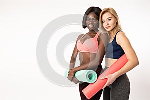 Pretty young african and european sportsgirls holding fitness mat looking at the camera while standing isolated over