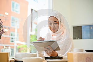 Pretty young asian muslim female online shop owner replying her customer question via tablet