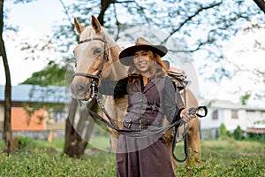 Pretty young Asian woman with cowboy costume smile and stand in front of brown horse and also look at camera in field near village