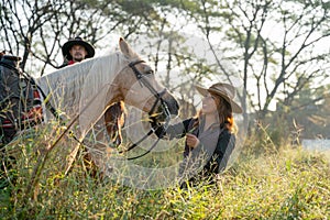 Pretty young Asian woman with cowboy costume smile and stand in front of white horse and stay near other cowboy man in field near