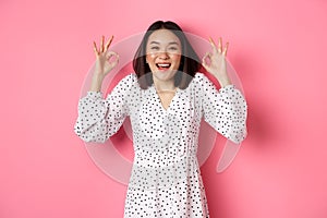 Pretty young asian woman in dress showing okay sign, praising and showing approval, looking satisfied, standing against