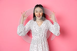 Pretty young asian woman in dress showing okay sign, praising and showing approval, looking satisfied, standing against