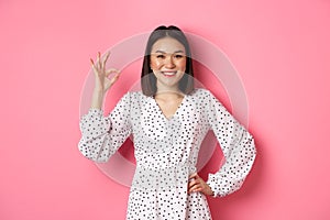 Pretty young asian woman in dress showing okay sign, praising and showing approval, looking satisfied, standing against