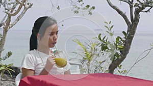 Pretty young Asian woman drinking pineapple juice on table near tropical seaside