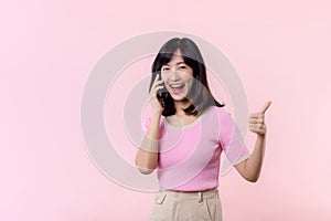 Pretty young asian woman showing success, victory hand gesture while receiving great news from smartphone on pink background.