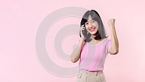 Pretty young asian woman showing success, victory hand gesture while receiving great news from smartphone on pink background.