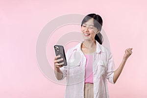 Pretty young asian woman showing success, victory hand gesture while receiving great news from smartphone on pink background.