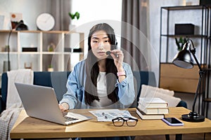 Pretty young Asian woman using headset and laptop for video call, working at home