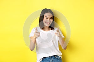 Pretty young asian woman in white t-shirt, pointing fingers at camera and smiling, praising or choosing you, say