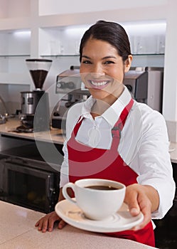 Pretty young barista offering cup of coffee smiling at camera