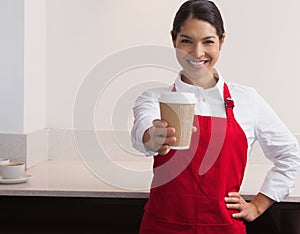 Pretty young barista offering cup of coffee to go smiling at camera