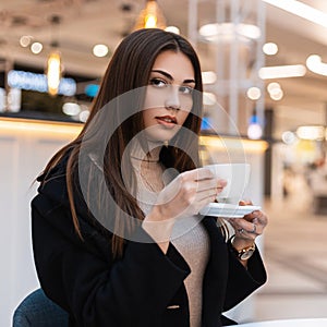 Pretty young beautiful brunette woman with long hair in black fashionable coat with cup of coffee is sitting in cafe in mall.