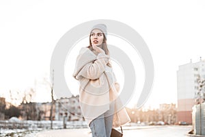 Pretty young beautiful woman in a fashionable knitted hat in a stylish eco coat of milk color with a leather handbag enjoys bright