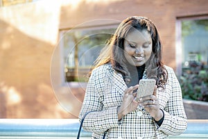 Pretty young black girl smiling writing sms on mobile phone outside office building with unfocused background