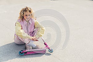 Pretty young blond curly haired hipster woman sits on the ground smiling, legs crossed, pink scooter in front of her
