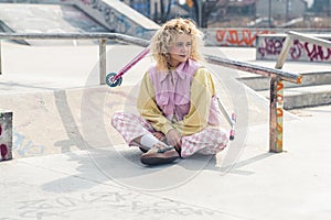 Pretty young blond curly haired woman sitting on the grey ground and looking away, smiling, legs crossed, a scooter on