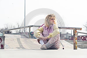 Pretty young blond ruddy hipster woman sits on the skateboard ground smiling, legs crossed, looking away pink scooter on