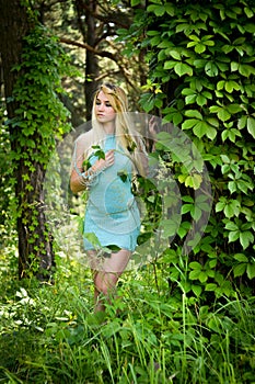 Pretty young blonde girl with closed eyes and long hair in turquoise dress standing in the green forest where trees are enlaced wi