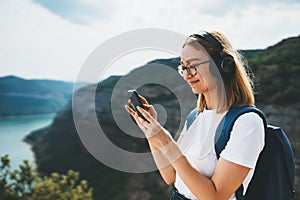 Pretty young blonde girl with glasses and backpack listens to her favorite music in wireless headphones and uses smartphone while