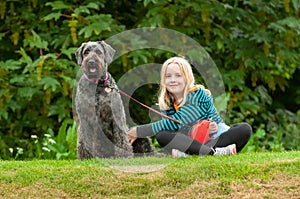 Pretty young blonde girl making eye contact while sitting on grass with well behaved black labradoodle dog on a leash
