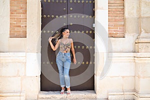 Pretty young blonde Spanish woman touches her hair, while looking to the side leaning against a wooden door. The woman is dressed