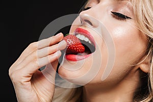 Pretty young blonde woman with bright makeup red lips posing isolated over black wall background eat strawberry
